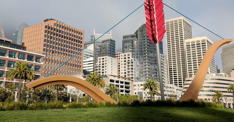 The giant Cupid's Arrow art installation in SF, in dappled sunlight in front of a cityscape with a long hanging fog over it.