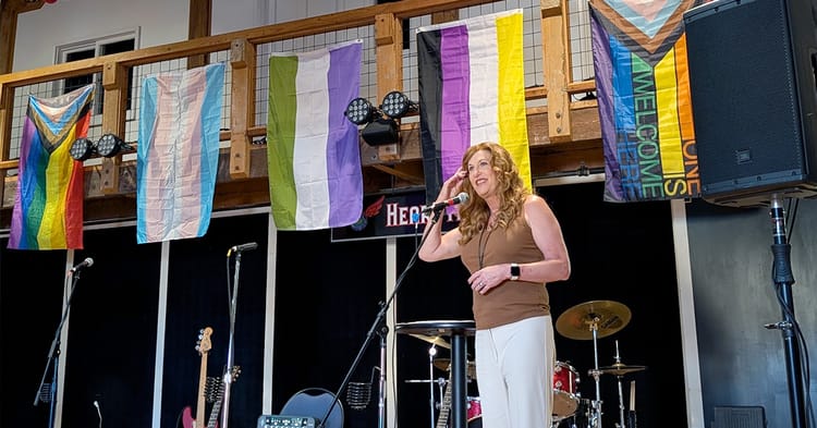 A photo of Suzanne Ford at the microphone, flanked by the band's instruments and speakers, with a variety of pride flags hanging from the banisters behind her.