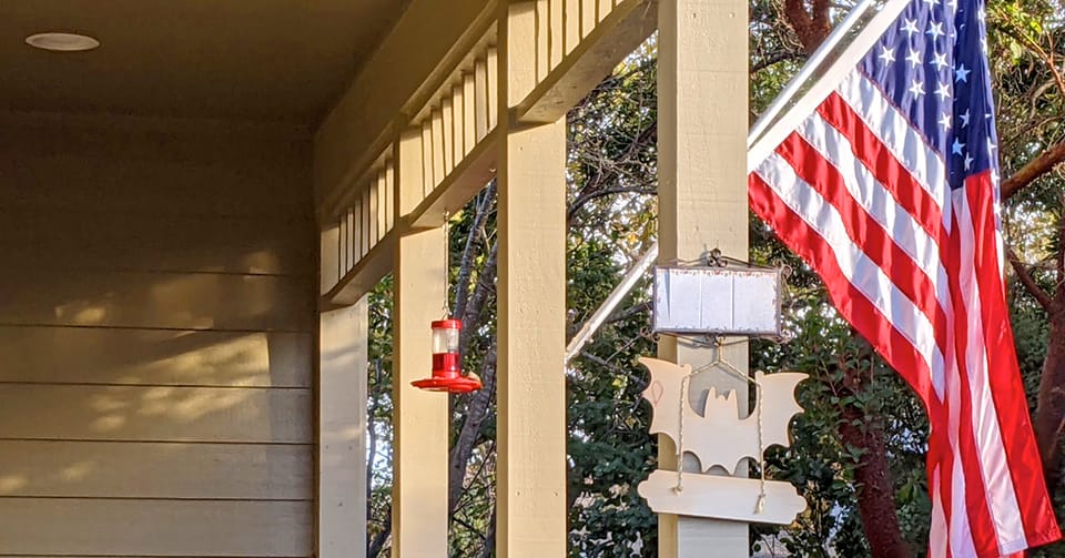 A photo of a home's porch, with the US flag flying near some Halloween decor and a hummingbird bird feeder.